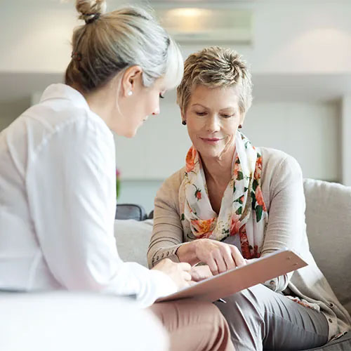 Two women sit together on a couch, one holding a clipboard and pen while the other points at the paper, both engaged in conversation in a bright, comfortable setting.