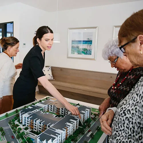 A woman points at a scale model of a modern apartment complex while three older women look on attentively in a well-lit room with framed art on the wall.