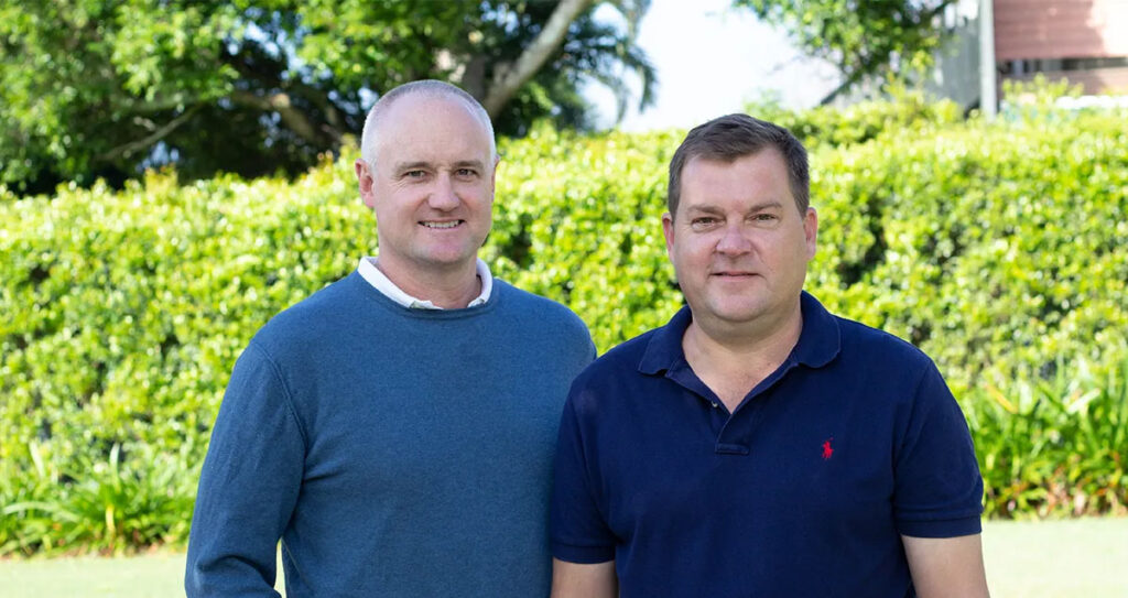 Two men standing side by side outdoors in front of a green hedge. One wears a blue sweater over a collared shirt, and the other wears a navy polo shirt. There are trees and a building in the background.