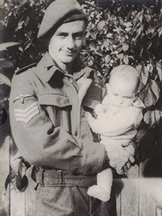 A black and white photo of a soldier in uniform holding a baby. The soldier is wearing a beret and standing in front of a leafy background. The baby is dressed in light-colored clothing.
