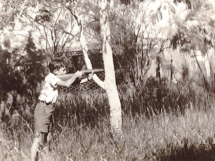 A black and white photo of a young boy wearing shorts and a shirt, aiming a rifle while standing near a tree. The background features tall grass and a fenced area with trees.