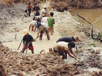 A group of people working in a muddy field near a river. They are using tools to dig or shape the earth. Some are wearing hats, and the scene suggests intense manual labor in a rural setting.