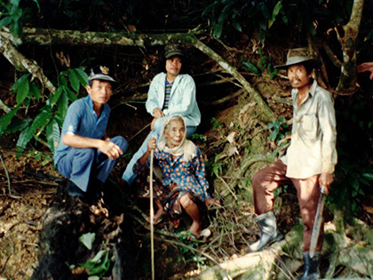 Four people are in a forest setting. Three men stand or squat; one man is in a blue outfit, another wears a hat and casual clothes, and the third man stands wearing brown. An elderly woman sits on the ground, holding a stick for support. Dense greenery surrounds them.