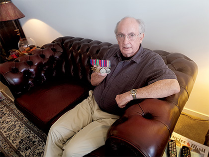 An elderly man sitting on a brown leather couch holds several military medals. He is wearing a gray shirt and beige pants. A lamp and remote controls are visible in the background.