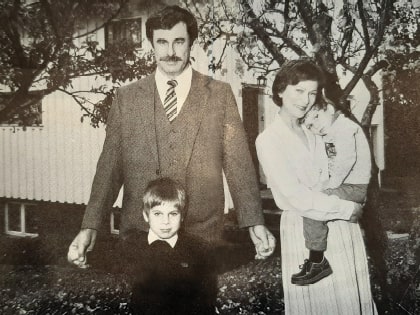 A black and white photo shows a man in a suit, a woman smiling while holding a child, and a young boy in front. They are standing outside in a garden with trees and a house in the background.