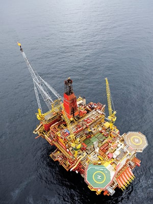 Aerial view of an offshore oil platform surrounded by the sea. The platform is equipped with cranes and has marked helipads. Its complex structure includes various decks and equipment painted in red and yellow.