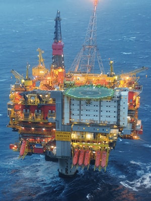 A large offshore oil drilling platform at sea, with numerous lights illuminating its structure. The platform includes a helipad and various cranes. The surrounding water is dark, suggesting evening or low-light conditions.