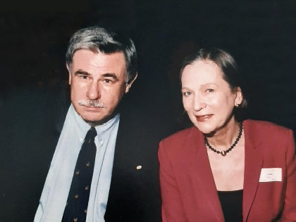 A man and woman are sitting and smiling. The man is wearing a suit and tie, and the woman is in a red blazer and necklace. They are seated closely together against a dark background.