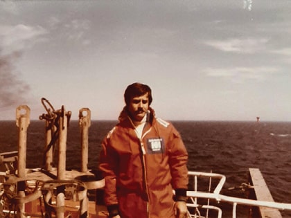 A man in an orange jacket stands on a ship's deck with the ocean in the background. He appears to be looking at the camera, and the sky above is clear with a few clouds. There is equipment visible on the deck near him.