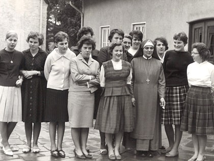 A group of women in vintage clothing stands together outside a building. One woman is dressed in a nun's habit. The others are wearing dresses and skirts typical of mid-20th century fashion. The setting appears to be a courtyard.