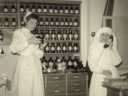 Two nurses in a vintage pharmacy setting. One nurse is holding a bottle and smiling, while the other is speaking on a telephone. Behind them are shelves filled with jars and bottles. They are wearing traditional nursing uniforms.