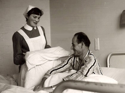 A black and white photo of a nurse in a uniform holding a pillow and smiling at a patient in a hospital bed. The patient, wearing striped pajamas, is sitting up and smiling back at the nurse.