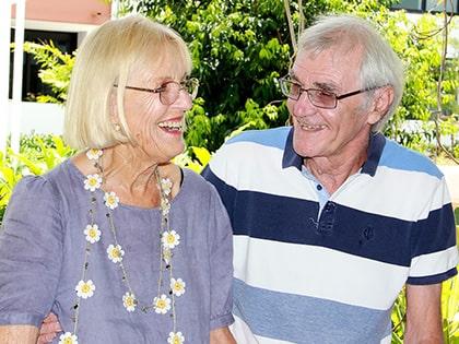 An elderly couple stands outdoors, smiling and looking at each other. The woman wears glasses, a floral necklace, and a grey blouse. The man wears glasses and a striped polo shirt. Greenery and buildings are visible in the background.