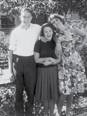 Three people pose happily outdoors in a black-and-white photo. A man in a white shirt stands on the left, with an arm around a woman in a dark outfit. Another woman in a floral dress stands on the right. Trees and a building are in the background.