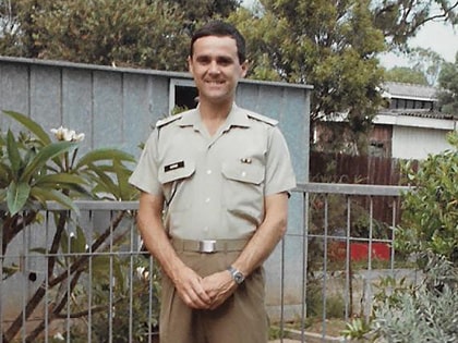 A man in a military uniform stands outside, smiling, with trees and a metal shed in the background. He is wearing a short-sleeved shirt with badges and brown trousers.