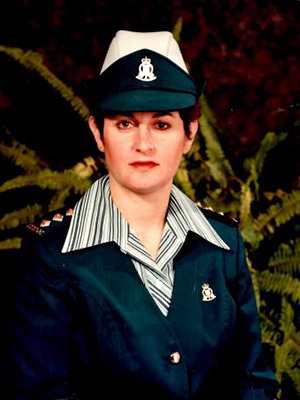 A person in a green military-style uniform with a matching hat and badges poses in front of a backdrop with ferns. They have short hair and a neutral expression.