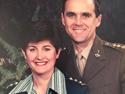 A smiling man and woman wearing military uniforms pose together in front of a dark background with green foliage.