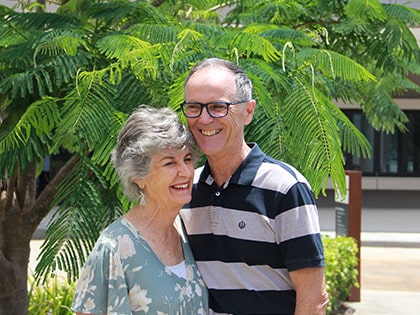 An elderly couple stands together, smiling happily under a tree with lush green leaves. The man wears glasses and a striped shirt, while the woman wears a floral blouse. They appear content and close, enjoying the outdoor setting.