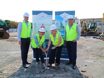 Four people in safety gear and wearing hard hats stand at a construction site. Two hold a shovel in the dirt. Banners in the background display building images and text. Construction equipment is visible around them.