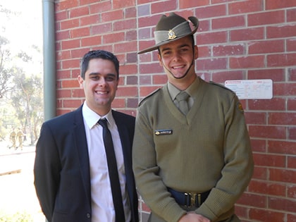 Two men are standing in front of a brick wall. One is wearing a suit and tie, and the other is in a military uniform with a khaki hat. They are both smiling.