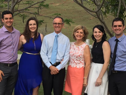 A group of six people, three men and three women, smile for a photo outdoors. They are dressed in formal attire with trees and a grassy field in the background.