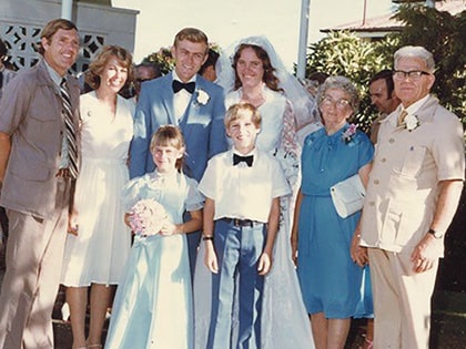 A wedding portrait featuring a bride in a white dress and veil, a groom in a blue suit, and several family members, including children in formal attire. They are posing outdoors, surrounded by greenery.