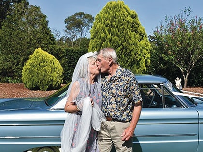 An elderly couple kisses in front of a vintage blue car. The woman is wearing a white veil, holding it in her hand. They are outdoors with lush greenery and trees in the background on a sunny day.