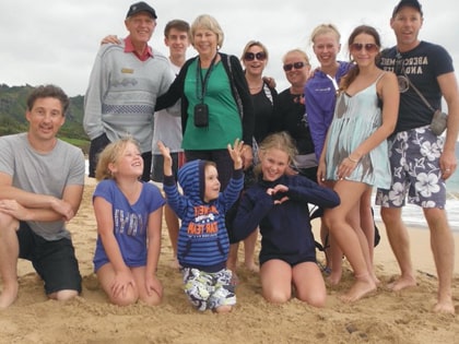 A group of people of various ages are gathered on a sandy beach, posing for a photo. Some are sitting on the sand while others stand behind them. The sky is overcast, and everyone is smiling.