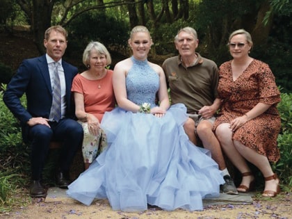 A family of five sits together on a park bench. The young woman in the center wears a light blue gown. The others are dressed in casual and semi-formal attire. Trees and greenery surround them, creating a serene outdoor setting.