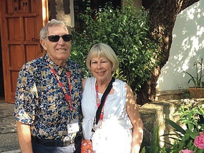 An older couple stands outside in a sunny garden. The man wears a floral shirt and sunglasses, while the woman is dressed in a white sleeveless top. Both smile warmly, surrounded by greenery and flowers, with a wooden door in the background.