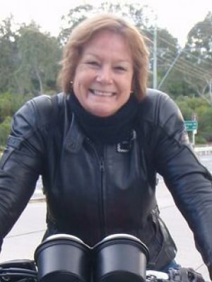 A person wearing a black leather jacket smiles while standing outdoors. They are leaning on what appears to be a motorcycle, with trees in the background.