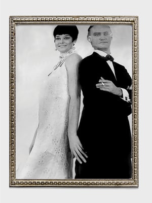A black and white photo of a woman in a sleeveless dress and a man in a suit and bow tie, both posing back to back. The image is in an ornate frame with a decorative border.