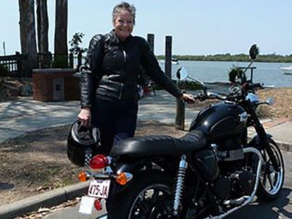 A person in a black leather jacket stands by a black motorcycle, holding a helmet. They are outdoors, near a body of water, with trees and parked boats in the background.