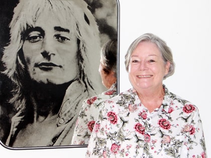 An older woman with short gray hair, wearing a floral blouse, smiles while standing next to a large black-and-white portrait of a person with long hair.