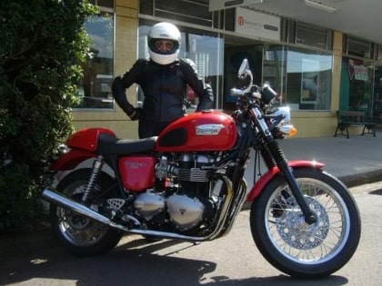 A person wearing a helmet and black motorcycle gear stands next to a red Triumph motorcycle parked on the street, with a building in the background.