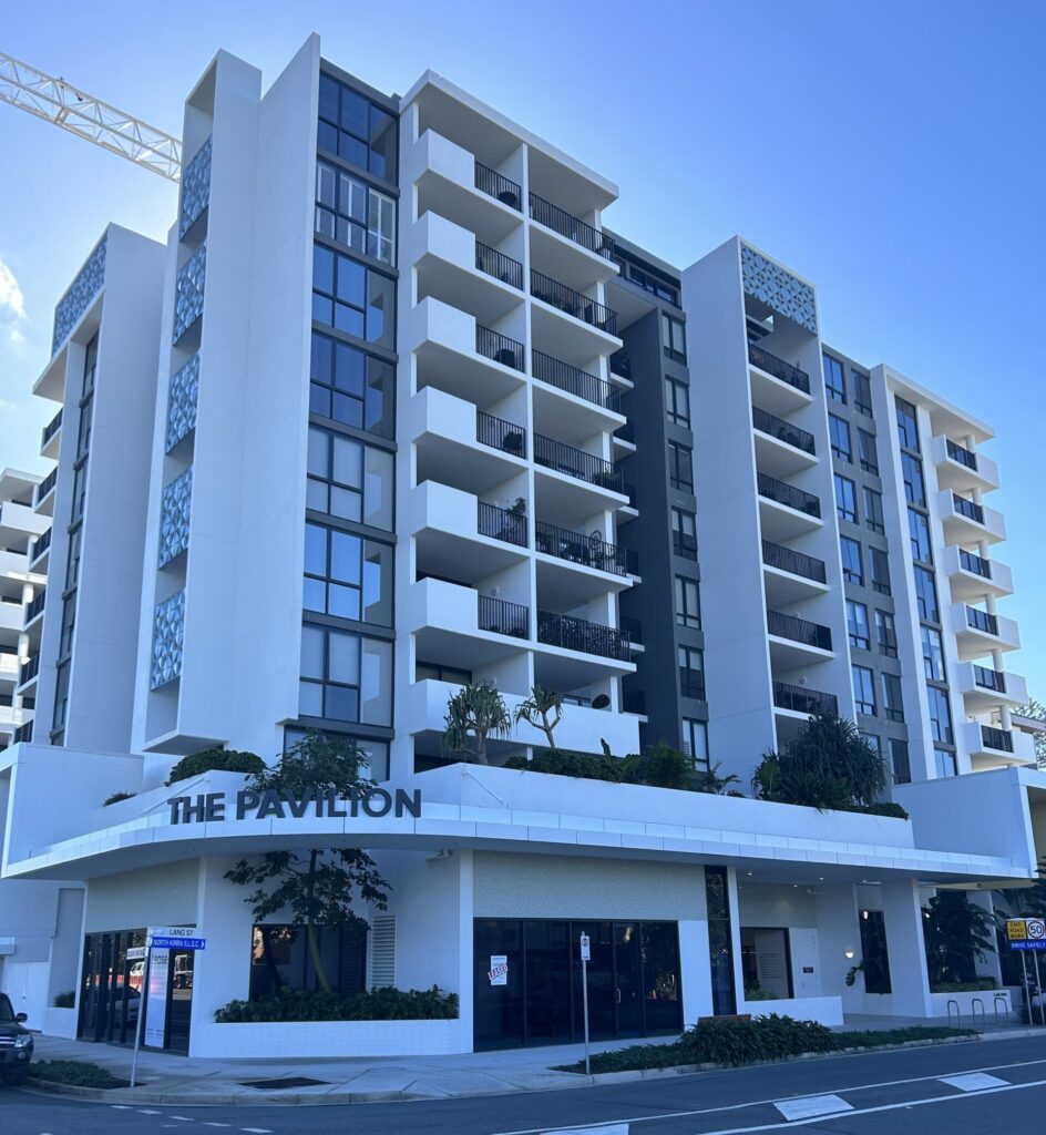 A modern multi-story apartment building named "The Pavilion" with large balconies and a sleek design. The structure features white and gray exterior walls, a ground-level commercial space, and landscaping around the entrance.