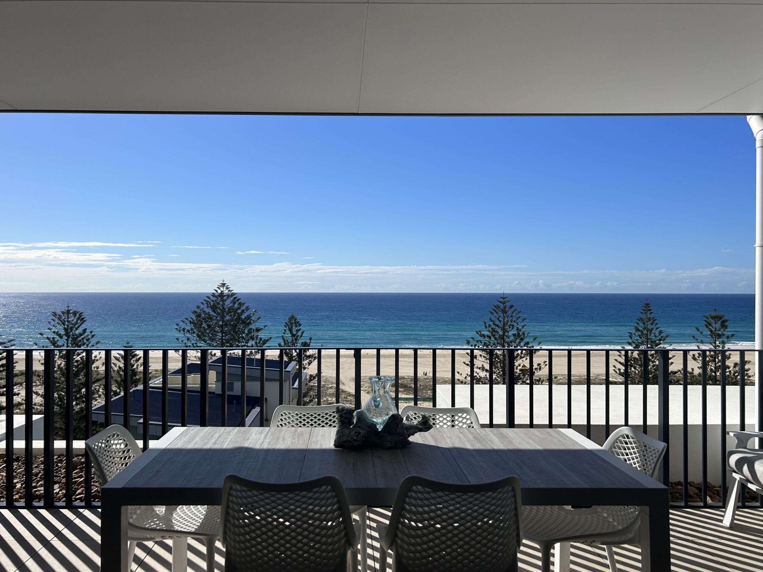 A modern balcony with a dining table and chairs overlooks a serene beach and ocean view. The clear blue sky and a few trees enhance the tranquil atmosphere.