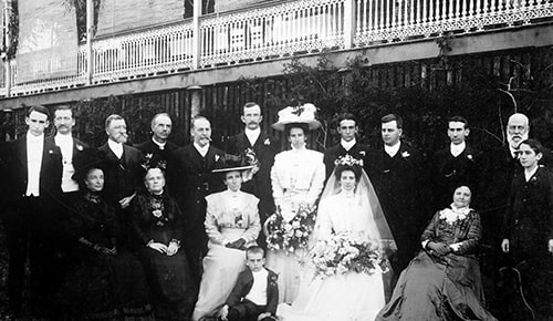 A vintage black and white photo of a large group dressed formally, likely at a wedding. The bride in a white gown and veil holds a bouquet, surrounded by family and friends, with men in suits and women in dresses, posed in front of a building.