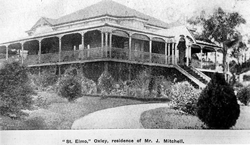 A black-and-white photo of a large, historic house with a wraparound porch and garden. It has a gabled roof and is elevated with a staircase leading to the entrance. Trees and shrubs surround the property.