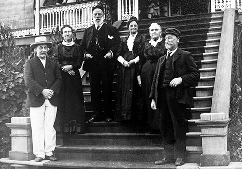 A group of six people, dressed in late 19th or early 20th-century attire, stand on the steps of a wooden house with a decorative railing. The men wear suits and hats, while the women are in long dresses with high collars.