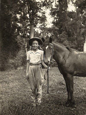 A vintage black and white photo of a person standing next to a horse in a grassy area. The person is wearing a hat, a collared shirt, and pants, holding the horse's reins. Tall trees are visible in the background.