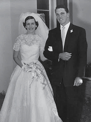 A black and white photo of a bride and groom. The bride is in a white lace dress holding a bouquet, and the groom is in a dark suit with a white tie. They are smiling and standing arm in arm in front of a doorway.