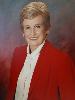 A person with short hair wearing a red blazer and white shirt smiles against a backdrop of blended colors.