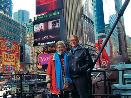 An older couple stands smiling in Times Square, New York City. They are surrounded by colorful billboards and advertisements, including Panasonic and Yahoo signs. The sky is clear and tall buildings are visible in the background.