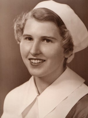 A sepia-toned portrait of a smiling woman in a nurse's uniform with a white cap. She has short, wavy hair and is facing slightly to the left. The image has a vintage feel.