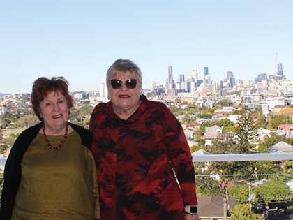 Two women smiling in front of a cityscape, featuring a clear blue sky and various skyscrapers. They are standing close together, with green and red clothing, on a balcony overlooking the city.