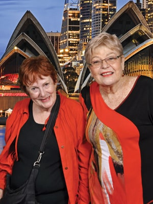Two older women smiling in front of a backdrop featuring the Sydney Opera House. One is wearing a red jacket and the other a red and black top. The evening sky and city lights create a vibrant background.