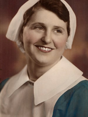 A woman in a historical nurse's uniform, with a white cap and collar over a blue dress, is smiling. The background is softly blurred in warm tones.