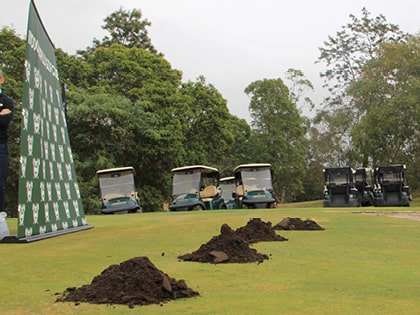 A row of golf carts is parked on a golf course near piles of soil on the grass. A green and white event backdrop stands to the side, and trees are visible in the background under an overcast sky.