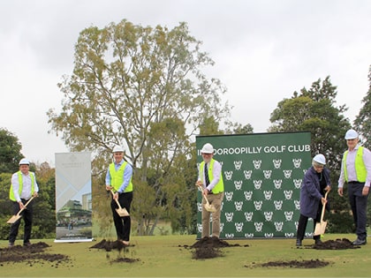 Five people in hard hats and safety vests break ground with shovels on a grassy area, with a banner reading "Indooroopilly Golf Club" in the background. Tall trees and other signs are also visible.